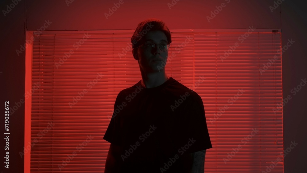 Portrait of male in dark room. Handsome man near window, red neon light shines behind jalousie, guy looks away naturally with calm expression.