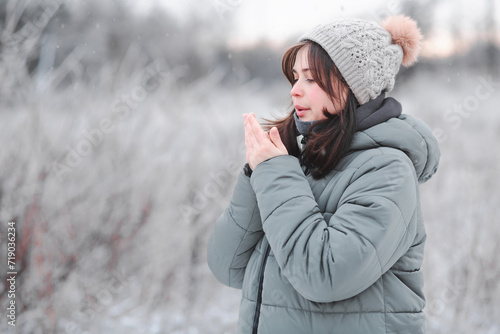 young woman in hat warms her hands with breath, cute girl walking on in frosty day, winter walk