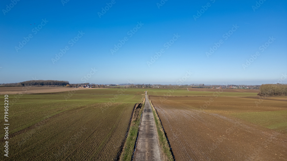 This aerial photograph offers a serene view of a straight country road as it divides expansive farmland under a clear blue sky. The fields on either side display varied stages of agricultural