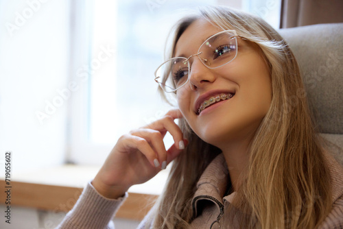 Close up portrait of young woman with dental braces system sitting in armchair near window and dreaming. Concept of beauty and medicine, dental care, malocclusion, orthodontic health. Ad photo