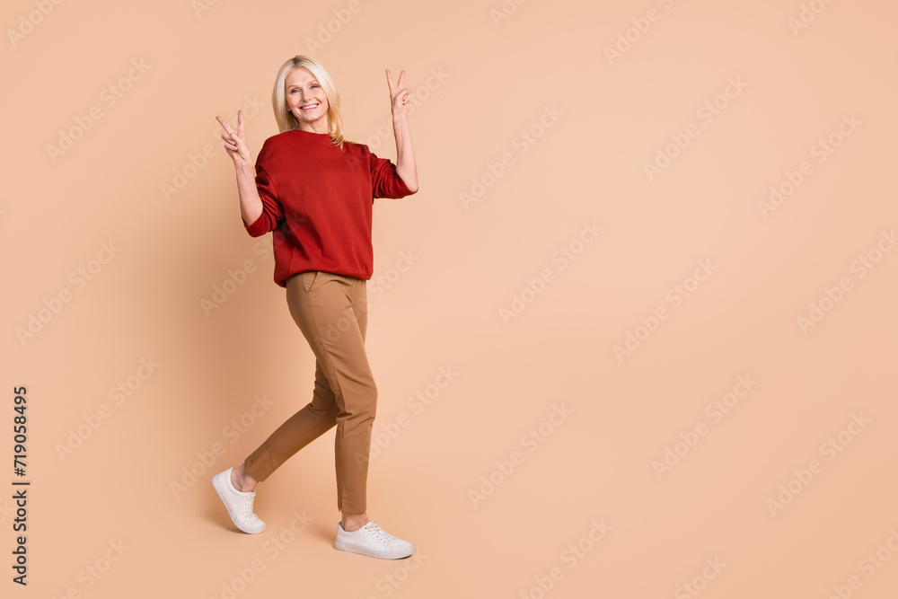 Full length photo of friendly funky lady dressed red pullover showing two v-signs walking empty space isolated beige color background Full length photo of friendly funky lady dressed red pullover showing two v-signs walking empty space isolated beige color background