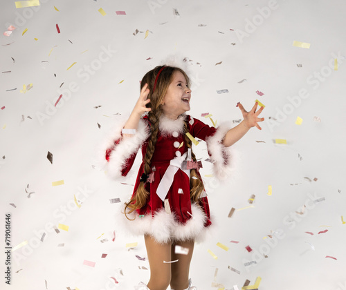 Happy Caucasian little girl wearing Christmas themed dress on gray studio background, copy space. Portrait of beautiful child having fun with sparkling confetti.