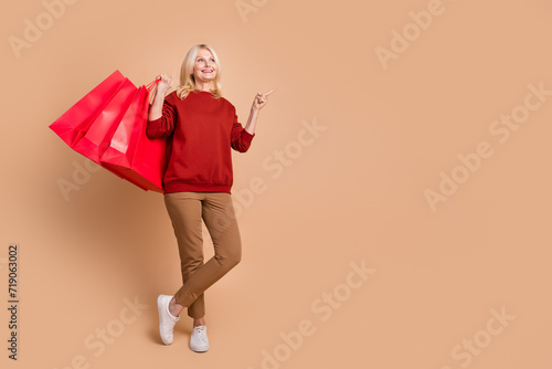 Full length photo of funky dreamy lady dressed red pullover holding shoppers pointing empty space isolated beige color background