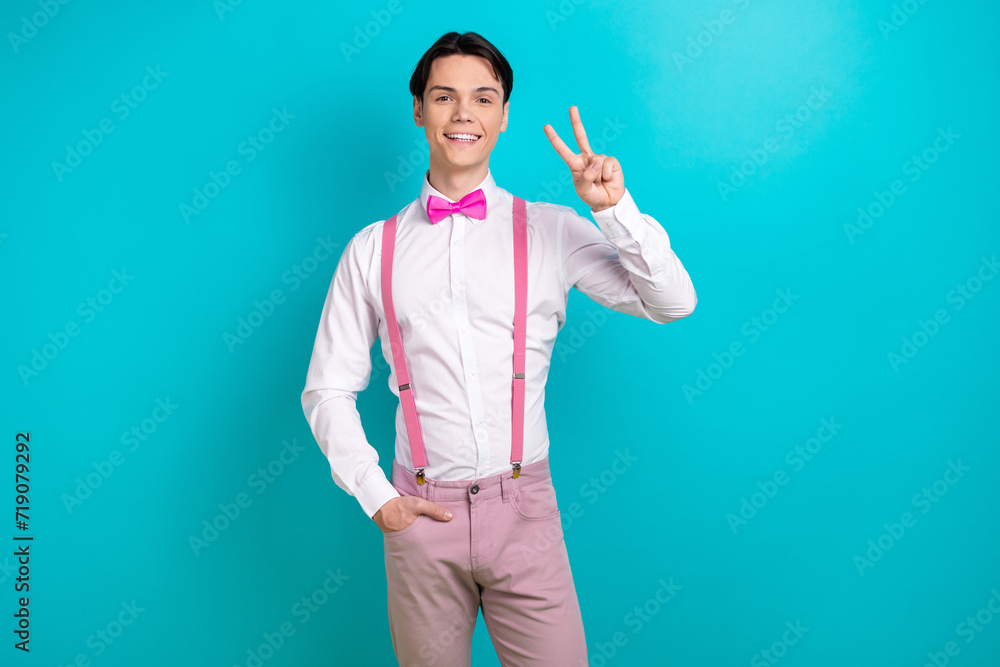 Photo portrait of pretty young male show v-sign greeting wear trendy pink outfit isolated on cyan color background Photo portrait of pretty young male show v-sign greeting wear trendy pink outfit isolated on cyan color background