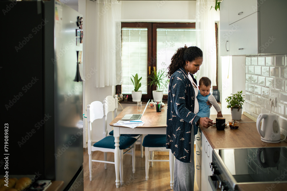 Mother holding baby while making coffee in a modern kitchen Mother holding baby while making coffee in a modern kitchen