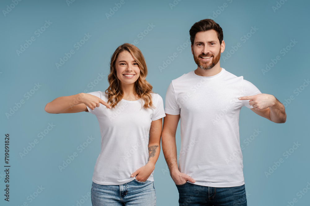Portrait of smiling confident couple, hipsters wearing white t shirt, pointing fingers isolated on blue background. Mockup concept Portrait of smiling confident couple, hipsters wearing white t shirt, pointing fingers isolated on blue background. Mockup concept