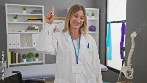 Joyful middle age blonde physio therapist strikes a thinking pose in rehab clinic, her confident face lighting up with a smart idea, as she pointedly raises one finger with a cheeky smile! photo