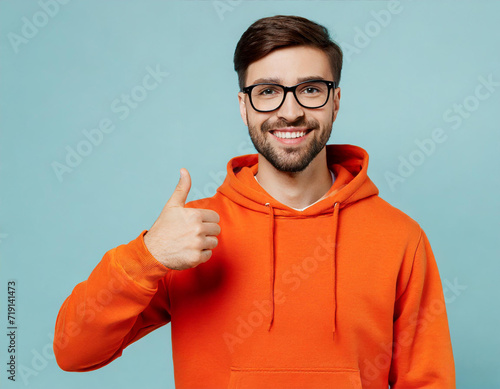 Young smiling happy fun man wear orange hoody casual clothes point thumb finger overhead on area mock up isolated on plain pastel light blue cyan color background studio portrait. Lifestyle concept