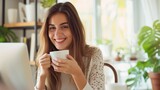 A happy emotionally inspired young woman looks into the camera holding cup of tea on home background .