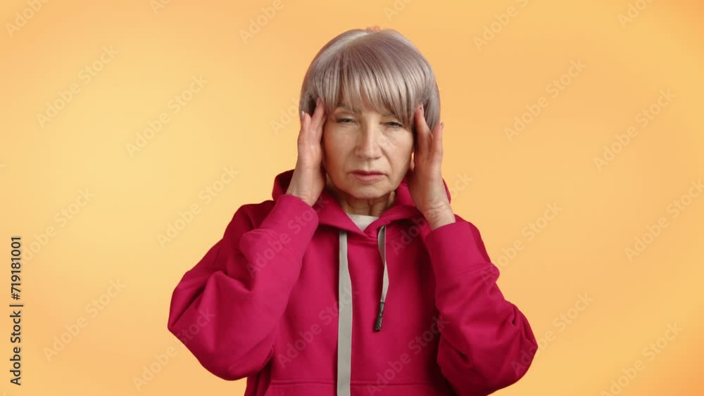 Elderly woman in a pink hoodie appears distressed, pressing her temples, indicating severe headache or migraine symptoms on a solid yellow background. Camera 8K RAW. 