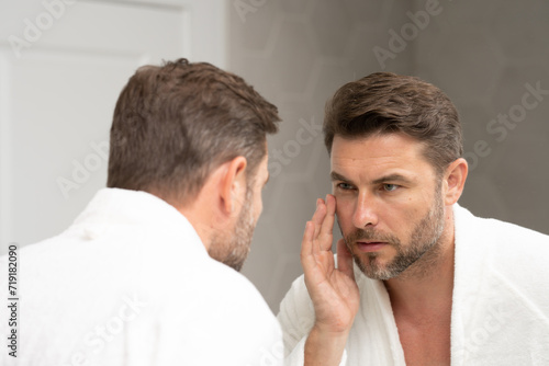 a man in a white bathrobe in the bathroom looking at the skin on his faceSpa male model, skincare product for skin face. Skin care. Male face cream. Man with perfect soft skin. Beauty cosmetic product photo