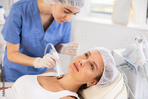 Young adult female client undergoing anti-aging ultrasonic procedure on neck at modern aesthetic medicine clinic for skin tightening and rejuvenation. Modern hardware cosmetology