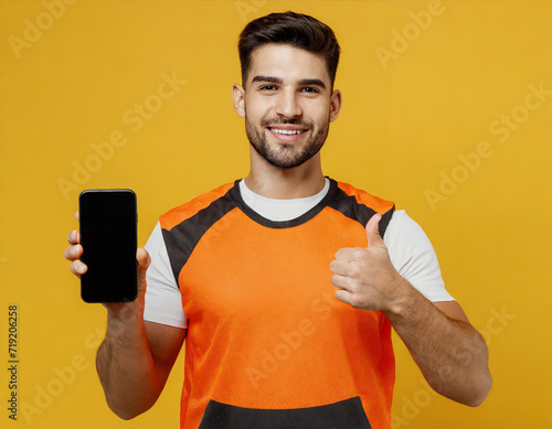 Young fitness trainer sporty man sportsman wear orange t-shirt hold use mobile cell phone show thumb up like spend time in home gym isolated on plain yellow background. Workout sport fit abs concept.