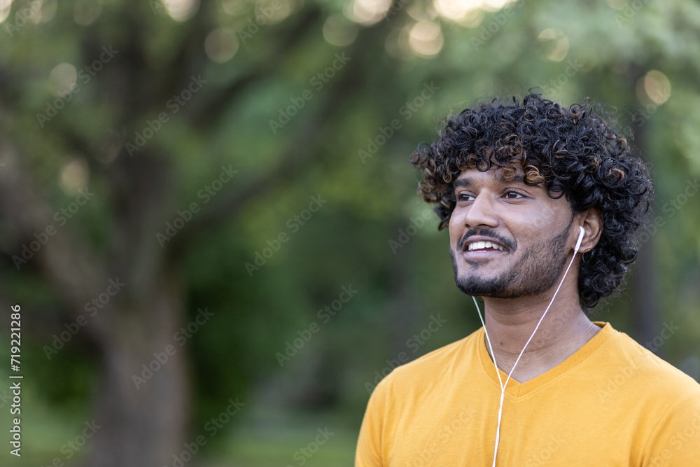 A young Indian man sportsman in headphones is standing in the park, smiling and looking to the side. Close-up photo A young Indian man sportsman in headphones is standing in the park, smiling and looking to the side. Close-up photo