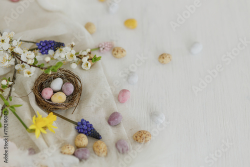 Colorful easter chocolate eggs in nest, spring flowers, feathers and linen border composition on white wooden table. Space for text. Happy Easter! Seasons greetings