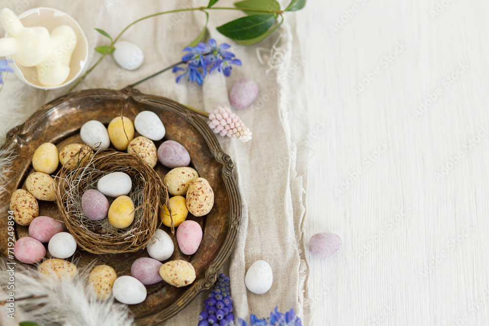 Happy Easter! Colorful easter chocolate eggs in nest, spring flowers, feathers and linen cloth on rustic wooden table. Space for text. Easter modern simple decoration