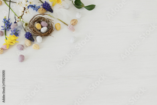 Happy Easter! Stylish easter chocolate eggs in nest, spring flowers, feathers border on white rustic wooden table. Easter modern simple decoration banner, space for text. Seasons greetings