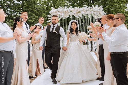 Bride and groom with friends near the ceremonial arch. Friends support the bride and groom. Lush and elegant bride's dress. Stylish wedding. Summer wedding in nature.