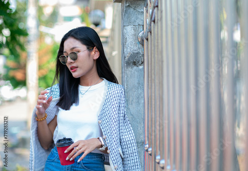A beautiful teenage girl wearing a white t-shirt, jeans and dark glasses holds a red teapot glass against the wall, looking cool and charming.