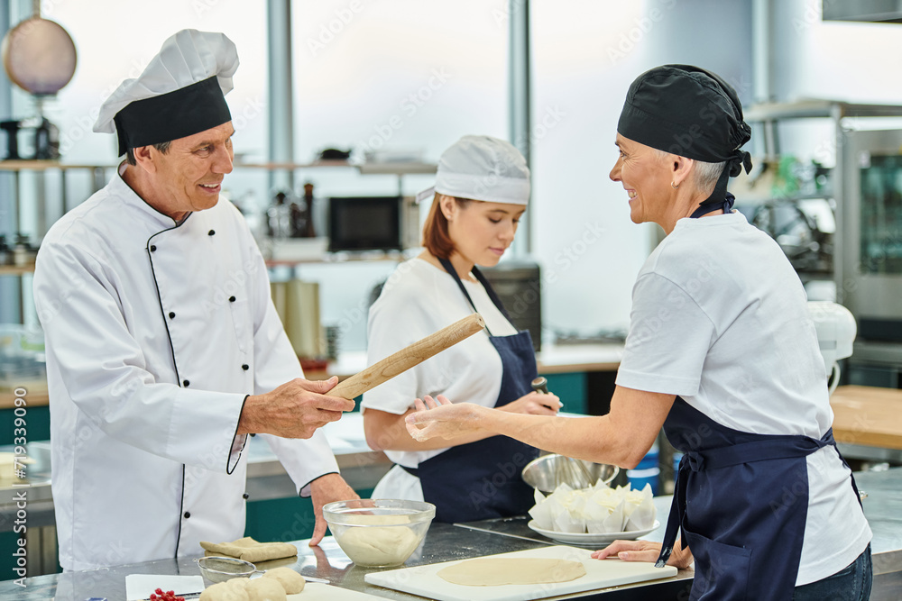 handsome cheerful chief cook giving rolling pin to his female chef next to their young colleague handsome cheerful chief cook giving rolling pin to his female chef next to their young colleague