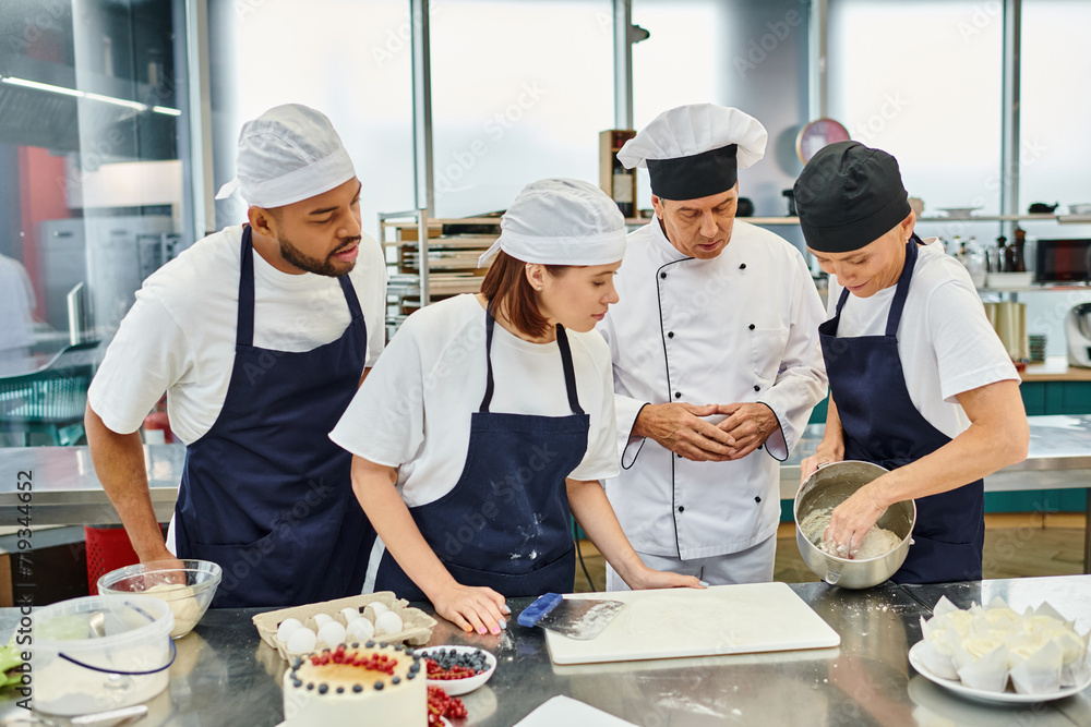 young man and woman in aprons watching with their chief cook how their colleague working with dough young man and woman in aprons watching with their chief cook how their colleague working with dough