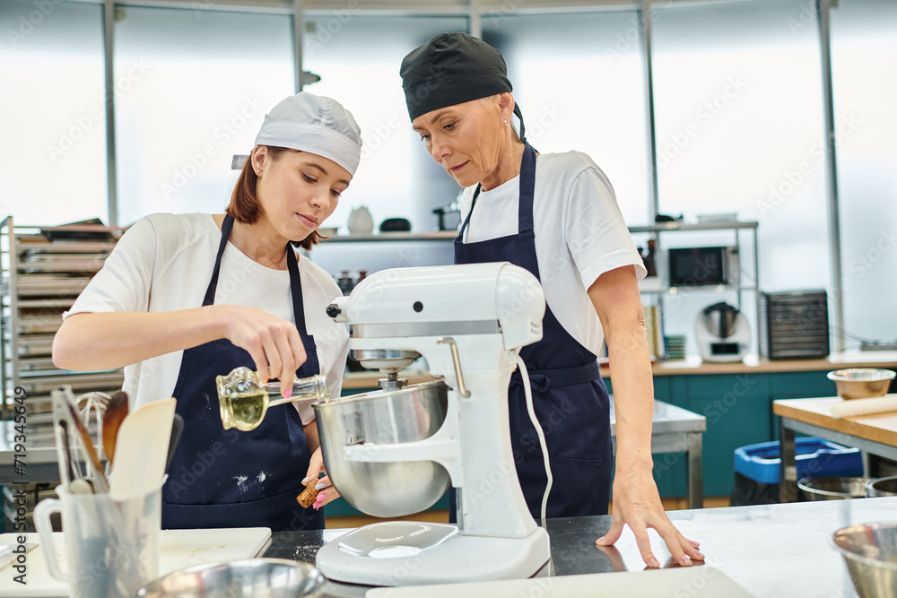 attractive female chefs in blue aprons and toques adding a little bit of oil to mixer, confectionery attractive female chefs in blue aprons and toques adding a little bit of oil to mixer, confectionery