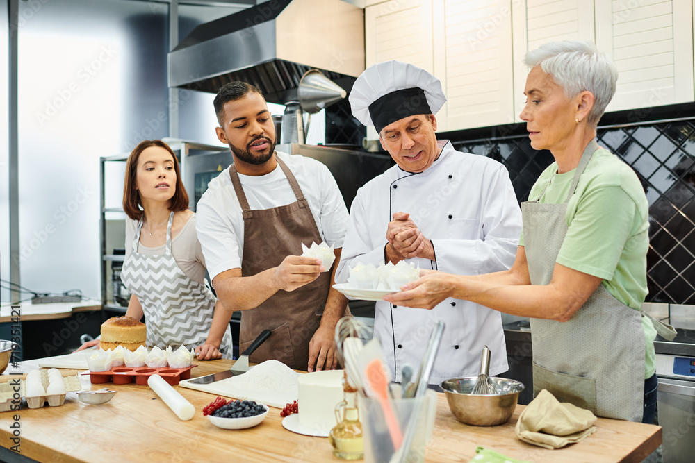 good looking interracial people in aprons listening to mature cheerful chef during cooking lesson - obrazy, fototapety, plakaty 