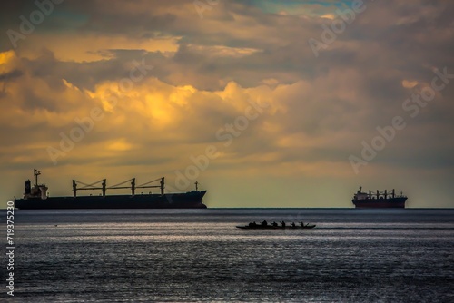 Silhouettes of two container ships contrasted with a silhouette of Indians paddling a long canoe, Vancouver harbor, Canada