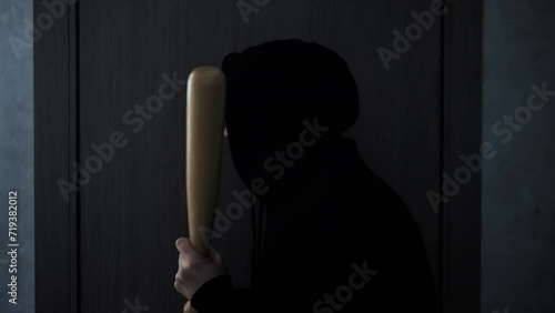 A man in a hood with a bat, robbing a house at night, aggressively knocks on a wooden door with his fist. Scandal with a knock on the door, a visit to the home of an aggressive man. photo