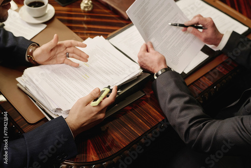 Two unrecognizable business partners discussing documents while sitting at wooden desk in boardroom