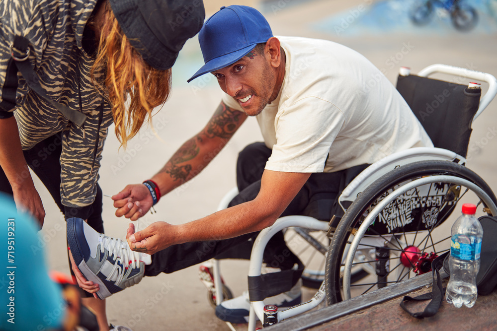 Obraz premium athlete man with disability in wheelchair smiling while fasten sneakers with help at skatepark 