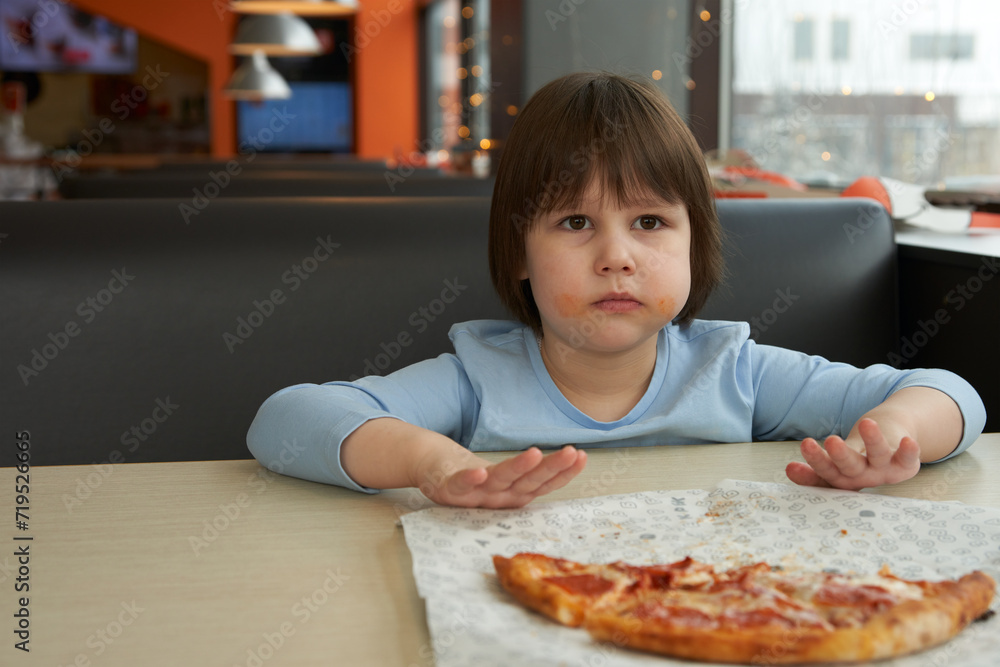 A child with his cheeks smeared with sauce pushes half a pizza away from him. The interior of a pizzeria in the background. Copy space. A child with his cheeks smeared with sauce pushes half a pizza away from him. The interior of a pizzeria in the background. Copy space.