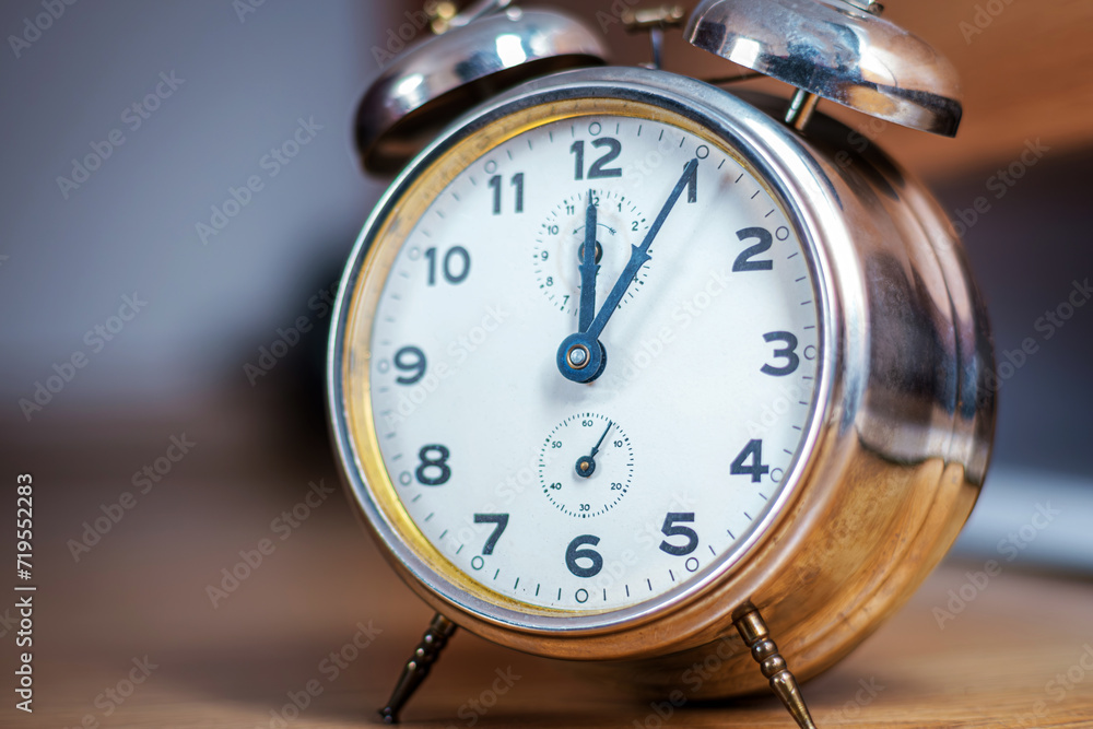 An aged metallic desk alarm clock. It shows noon. It stands out against the blurred background. Idea image about using time efficiently. Selective focus. An aged metallic desk alarm clock. It shows noon. It stands out against the blurred background. Idea image about using time efficiently. Selective focus.