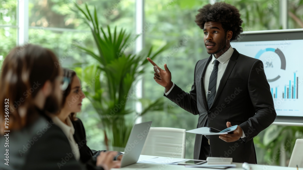 Business presentation taking place with a well-dressed man pointing at a chart on a display board while colleagues or clients listen attentively around a conference table. Business presentation taking place with a well-dressed man pointing at a chart on a display board while colleagues or clients listen attentively around a conference table.