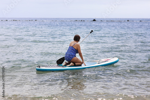 Middle aged woman in swimsuit sits on inflatable SUP board and paddling at calm ocean water. Active female enjoys does yoga, stretching or trains on paddle board at summer vacation. Healthy lifestyle © Andriy Medvediuk
