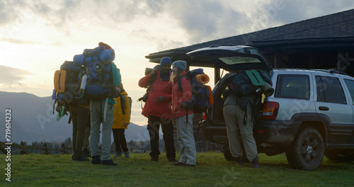 Full shot of diverse tourist friends arrive to rental house on hill. Happy hikers take luggage from trunk of car and talk. Hiking buddies during trip or expedition to mountains on their vacation.