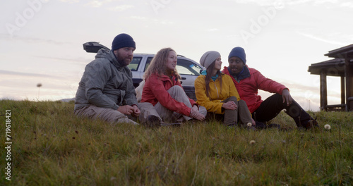Group of multiethnic tourists sit on the grass at the hilltop and talk. Travelers or hiking friends enjoy nature during trip to the mountains. Stylish wooden cottage, car and dog in the background.