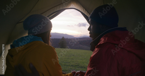 Multiethnic couple of hikers sits in tent on hilltop, kisses and looks at beautiful sunset. African American man and Caucasian woman enjoy amazing mountains scenery during trip. Outdoor enthusiasts.