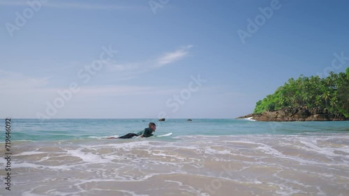 Male surfer in long sleeve rash guard paddling on surfboard. Man in rashguard swimwear swims to lineup lying on surfboard. Surfer goes to ocean water swells on his board. Recreational activity.