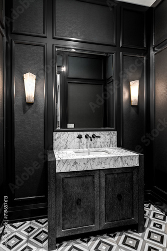 A bathroom with black wainscoting and wallpaper  a dark grey wood vanity cabinet  marble apron sink with black faucet  and mosaic tile flooring.