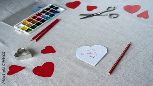 Valentine's day greeting card with red hearts, colorful paints, scissors and pencils on the table