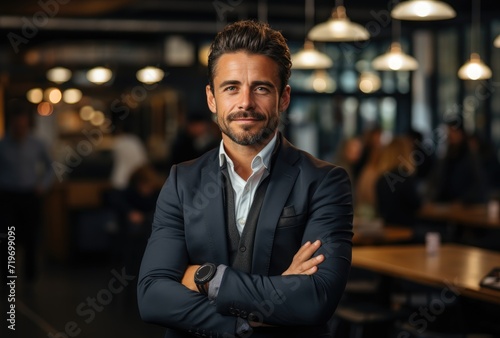 A sharply dressed businessman stands confidently with a charming smile, exuding professionalism and sophistication as he crosses his arms against the backdrop of an elegant indoor setting with a tabl