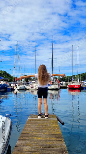 a young beautiful girl with long hair in a white shirt stands with her back to the viewer on a pier with yachts against the backdrop of the sea with her arms open, embracing the whole world and this w