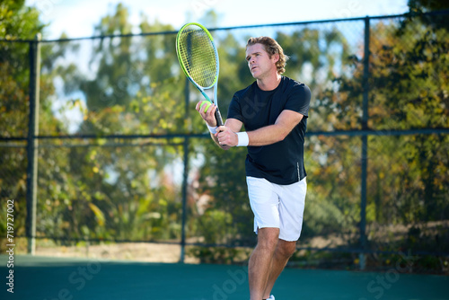 A man prepares to serve a tennis ball on a court surrounded by trees