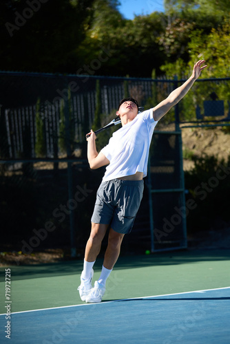 An asian man plays tennis on a court surrounded by trees