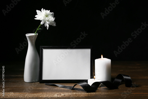 Blank funeral frame, burning candles and black ribbon on wooden table against dark background photo