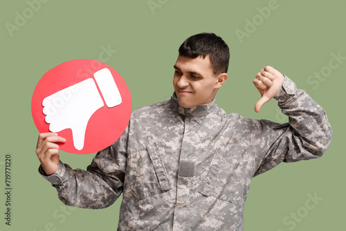 Young displeased male soldier with dislike icon showing thumb-down gesture on green background photo