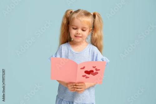 Cute little girl holding greeting card with hearts on blue background. Valentine's Day celebration