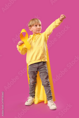 Cute little boy in superhero costume with yellow ribbon on purple background. Childhood cancer awareness concept