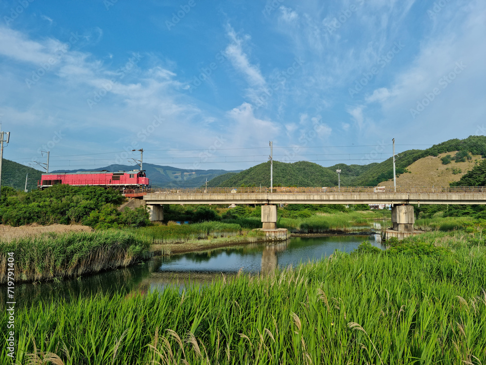 A rural scene with a red train crossing a bridge.